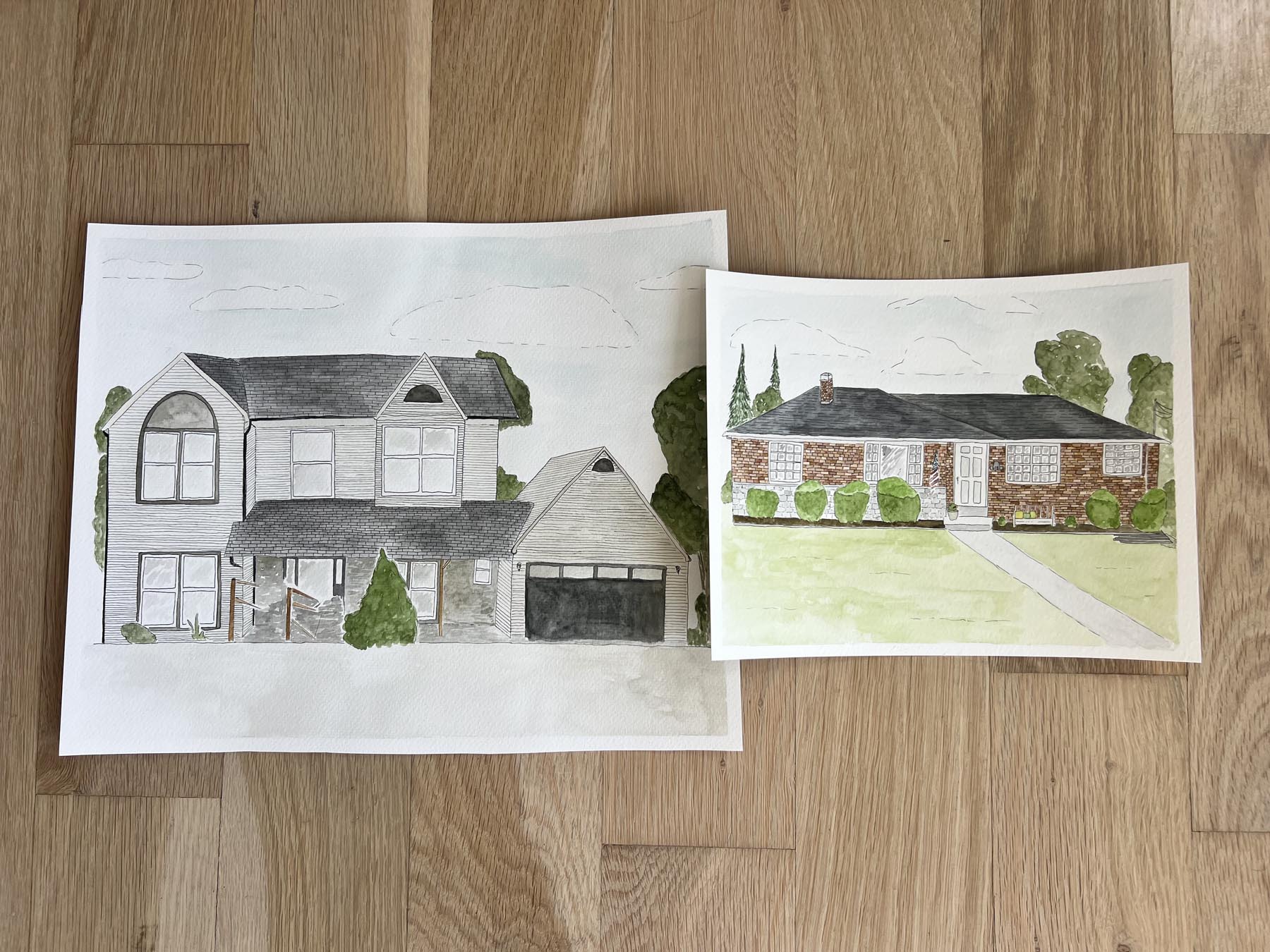 Two watercolor drawings of a house on a wooden surface.