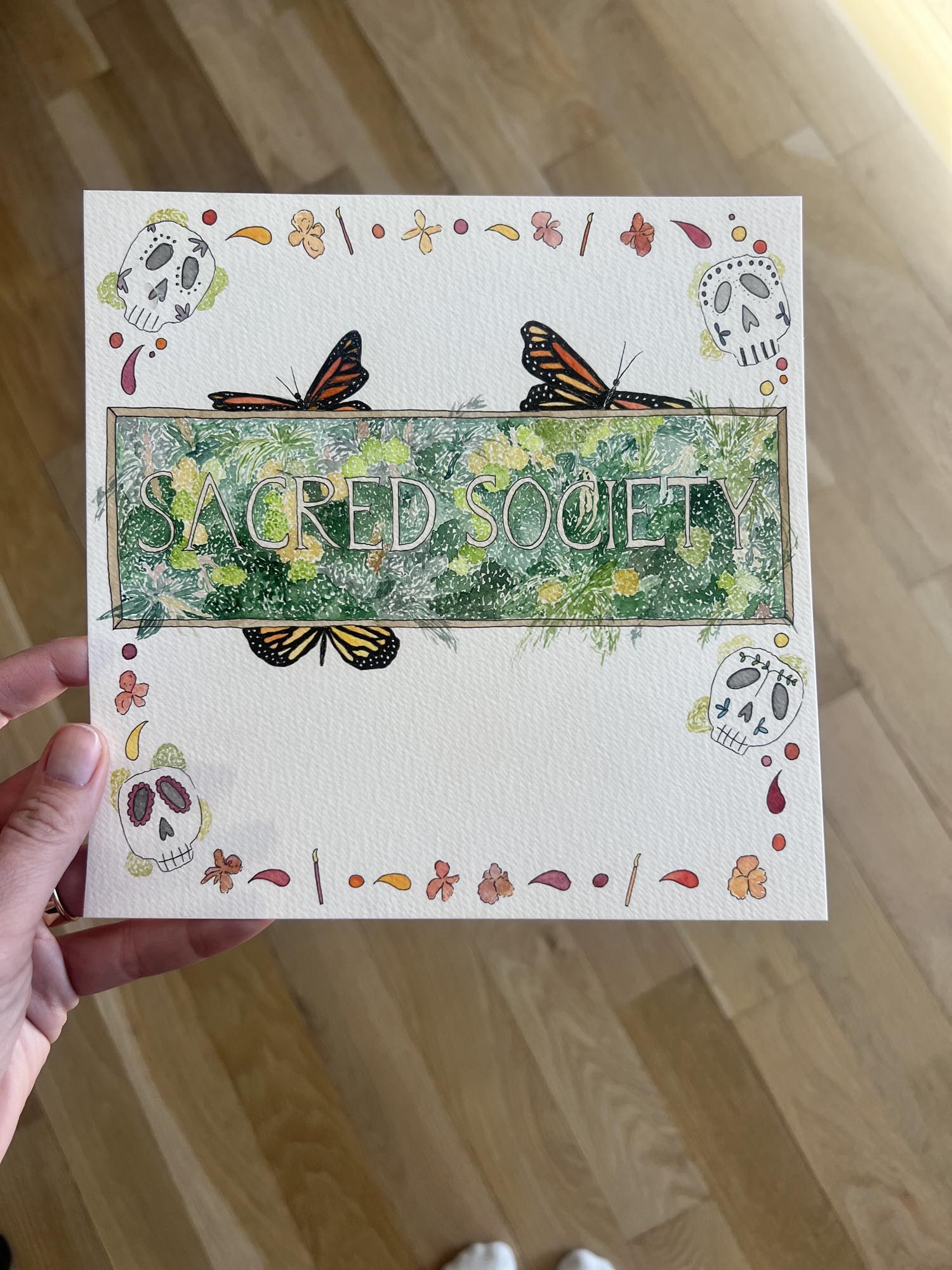 Hand holding a card with butterfly and nature designs and 'Sacred Society' text on a wooden floor.