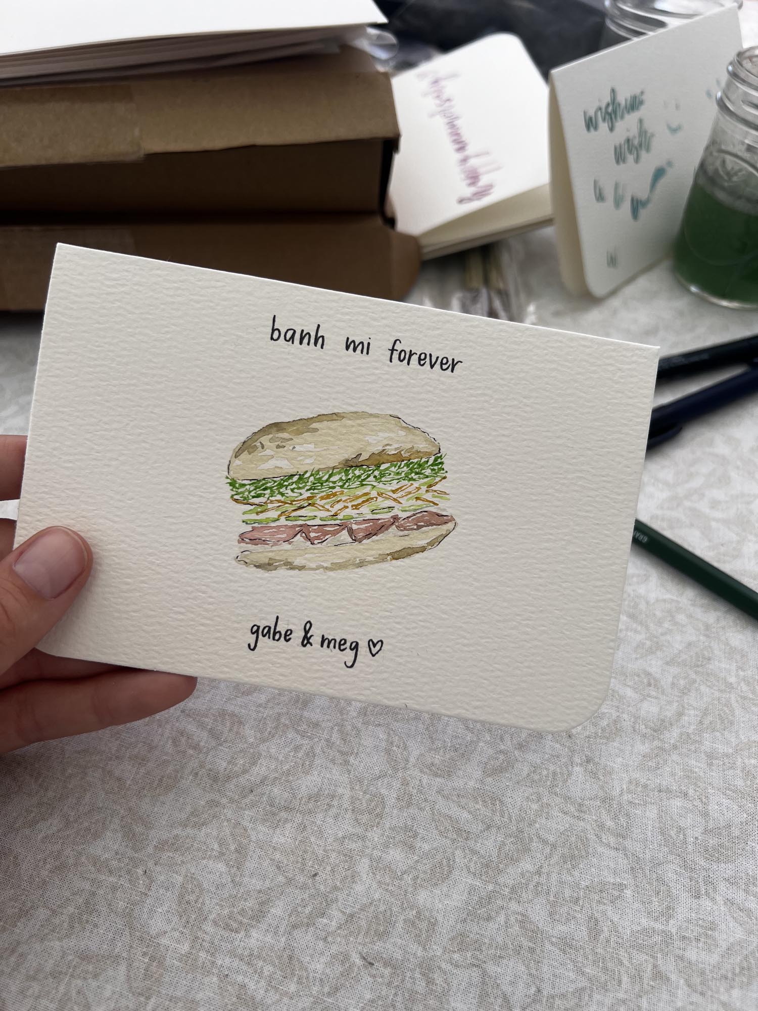 Card with a drawing of a banh mi sandwich and text 'banh mi forever gabe & meg' held by a hand.