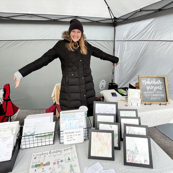 Woman standing behind a table with framed artwork and stationery items inside a tent.